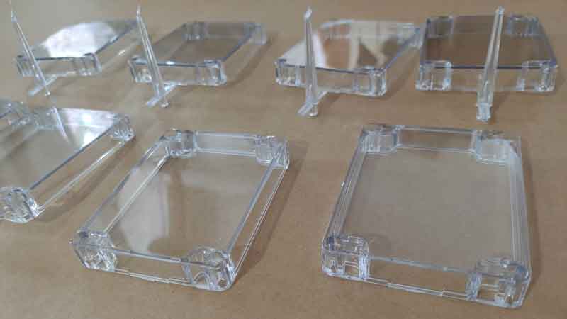 Clear plastic cover in production for a custom enclosure, with the sprue still attached, demonstrating quality control in the manufacturing process.